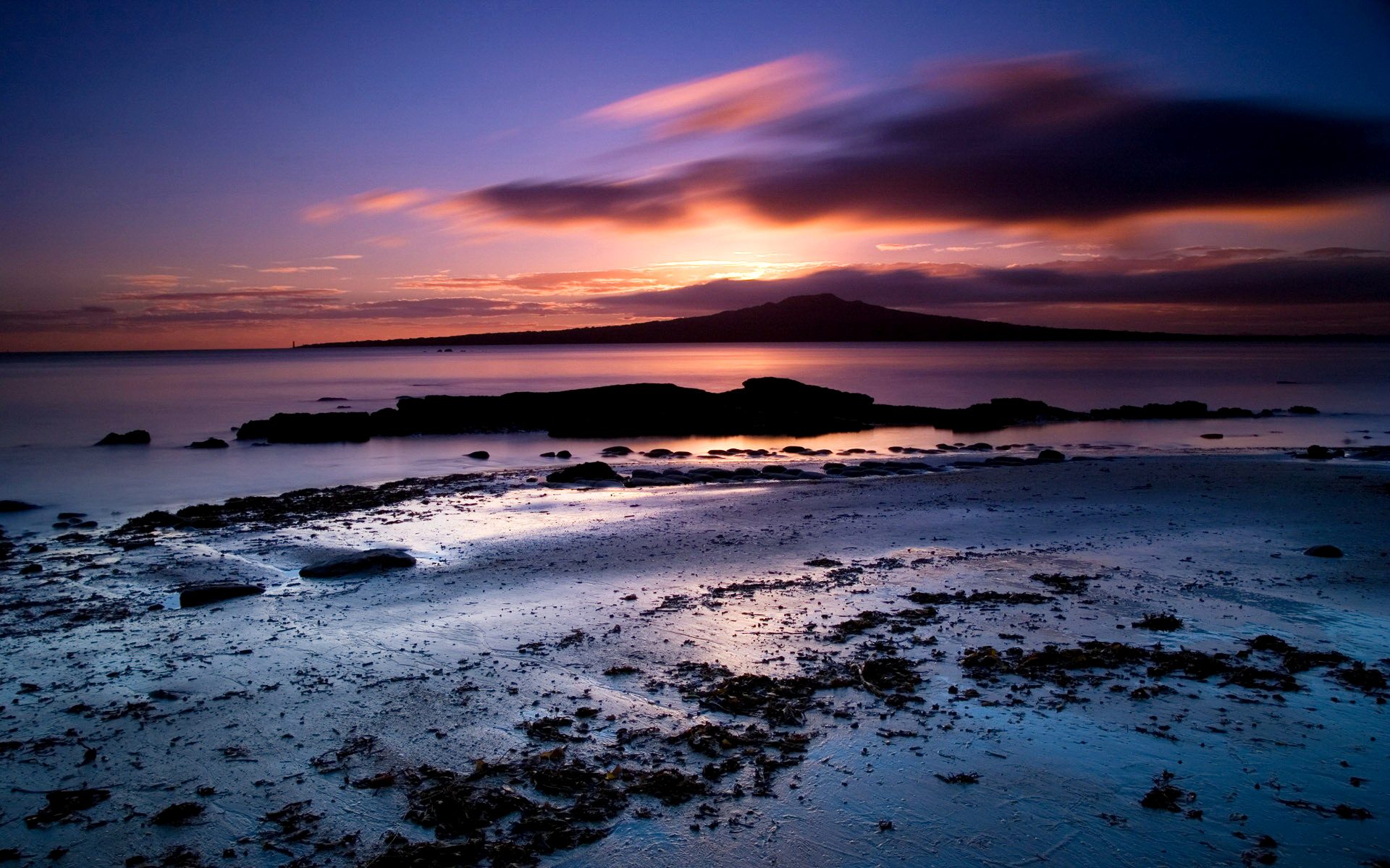 A breathtaking sunset over a tranquil beach, with the silhouette of Rangitoto Island in the distance, casting a warm glow on the wet sand and scattered seaweed