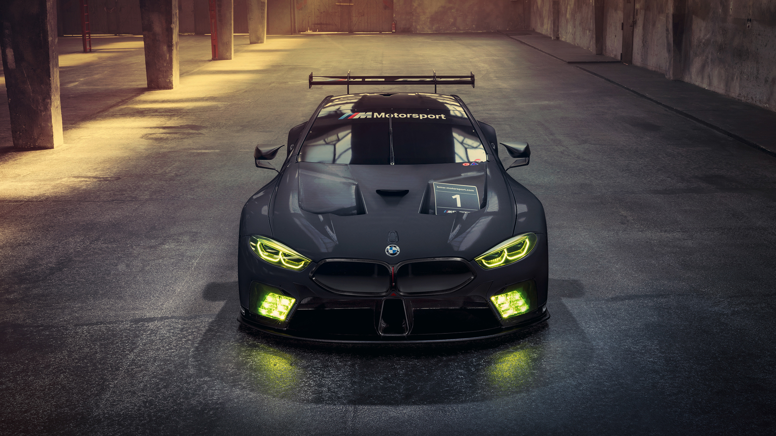 A front view of a sleek, black BMW M Motorsport race car, illuminated by its bright yellow headlights, parked in a dimly lit garage