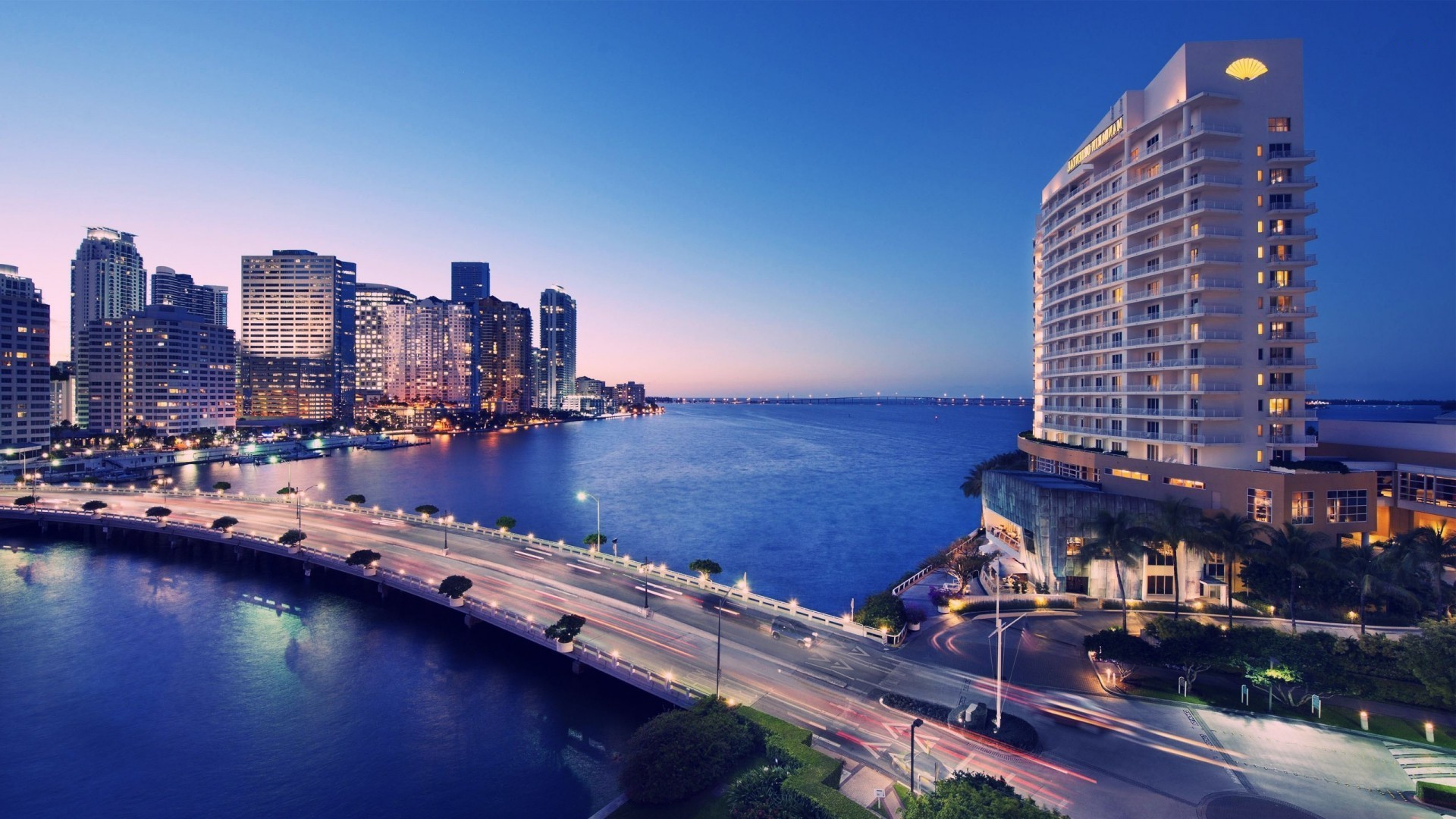 A stunning twilight view of the Miami skyline, featuring the illuminated cityscape, the Brickell Key Bridge with light trails, and a prominent waterfront hotel