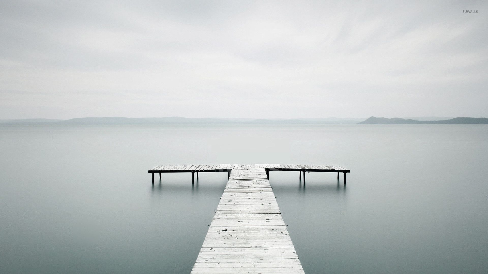 A minimalist photograph of a wooden pier extending into a calm, misty lake. The soft, muted colors and simple composition create a serene and peaceful atmosphere