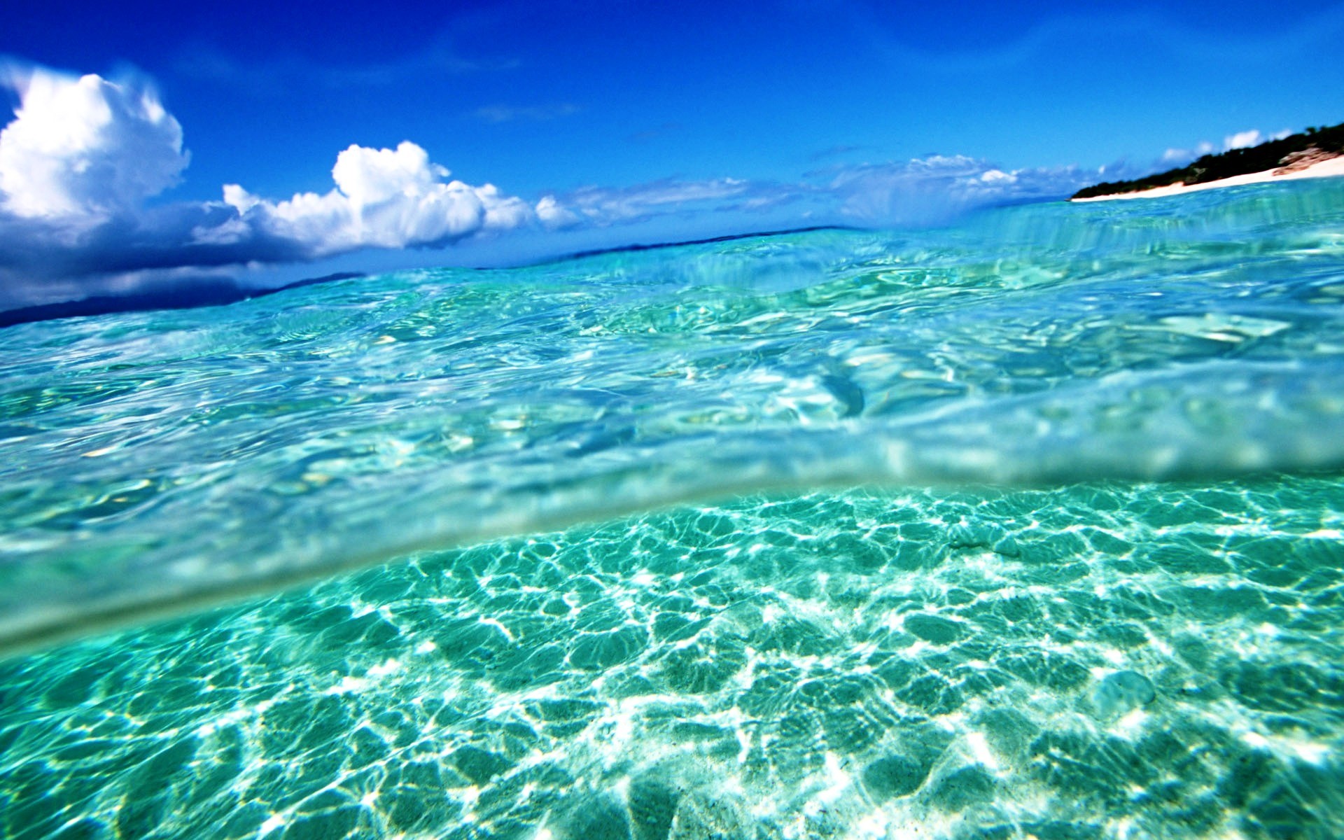 Crystal-clear turquoise water gently laps a white sand beach under a vibrant blue sky dotted with fluffy white clouds. This idyllic summer scene evokes a sense of tranquility and escape