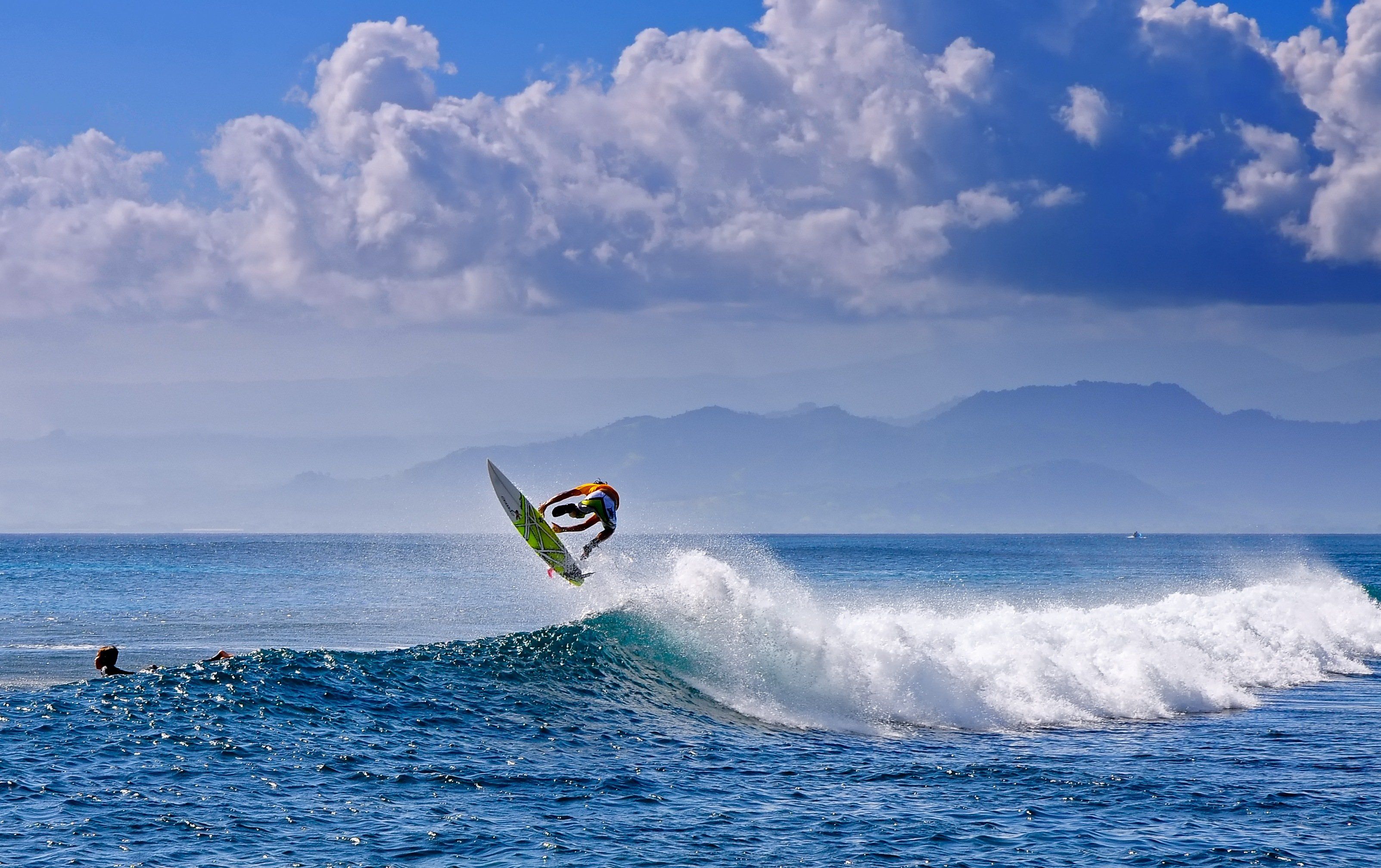 A surfer performs an aerial maneuver on a wave with a backdrop of mountains and a cloudy sky