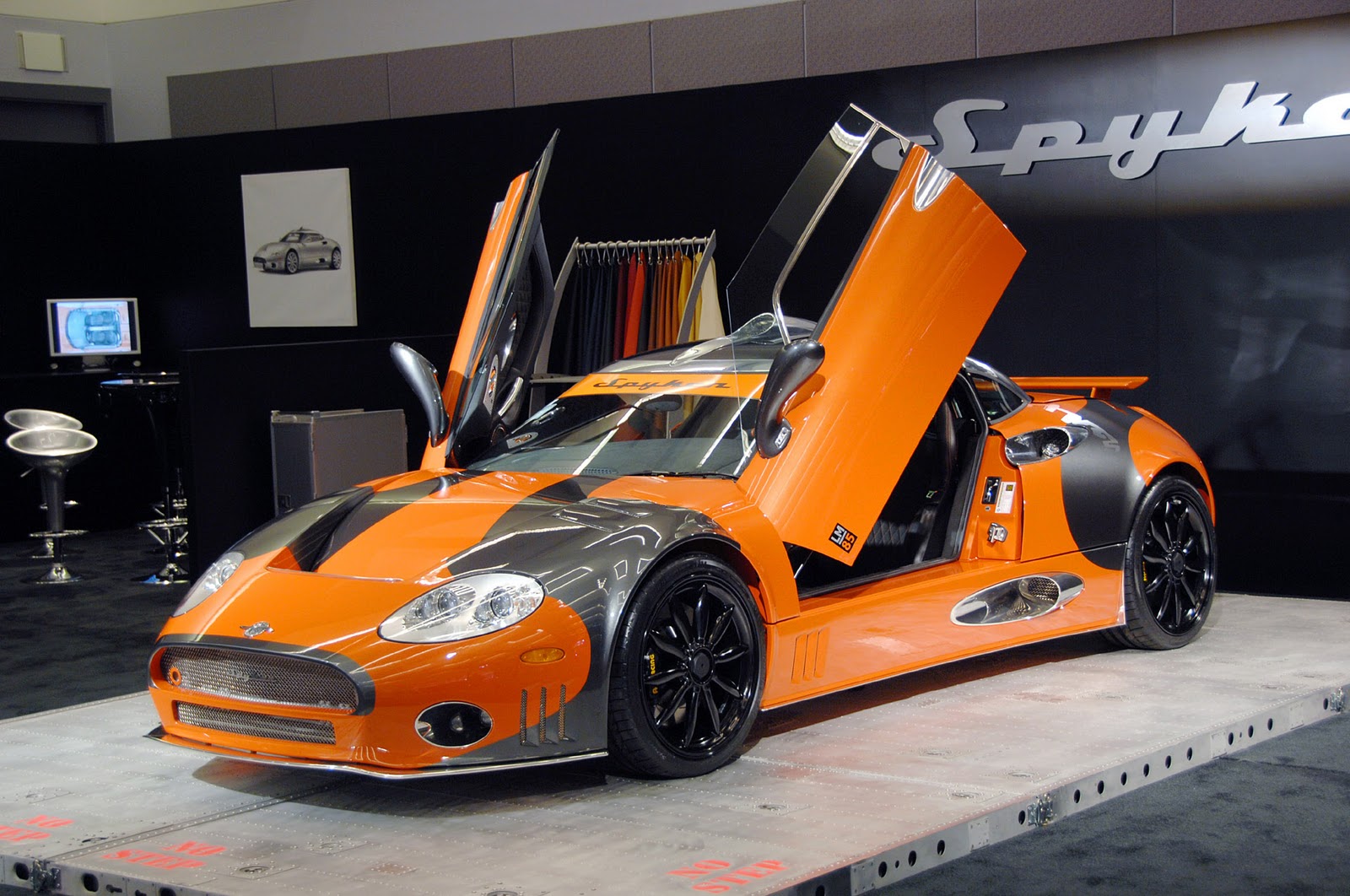 A vibrant orange and black Spyker C8 sports car with its distinctive scissor doors open, displayed at an auto show