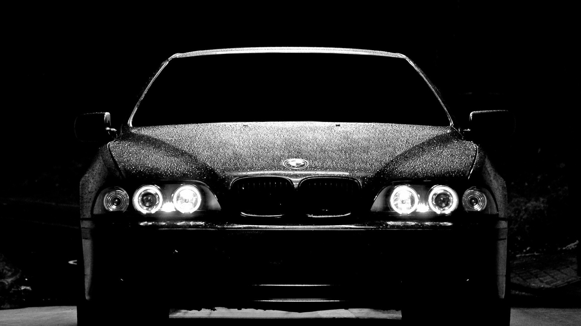 A sleek black BMW car, glistening with water droplets, parked in a dark setting with its headlights illuminated, exuding an aura of sophistication and power