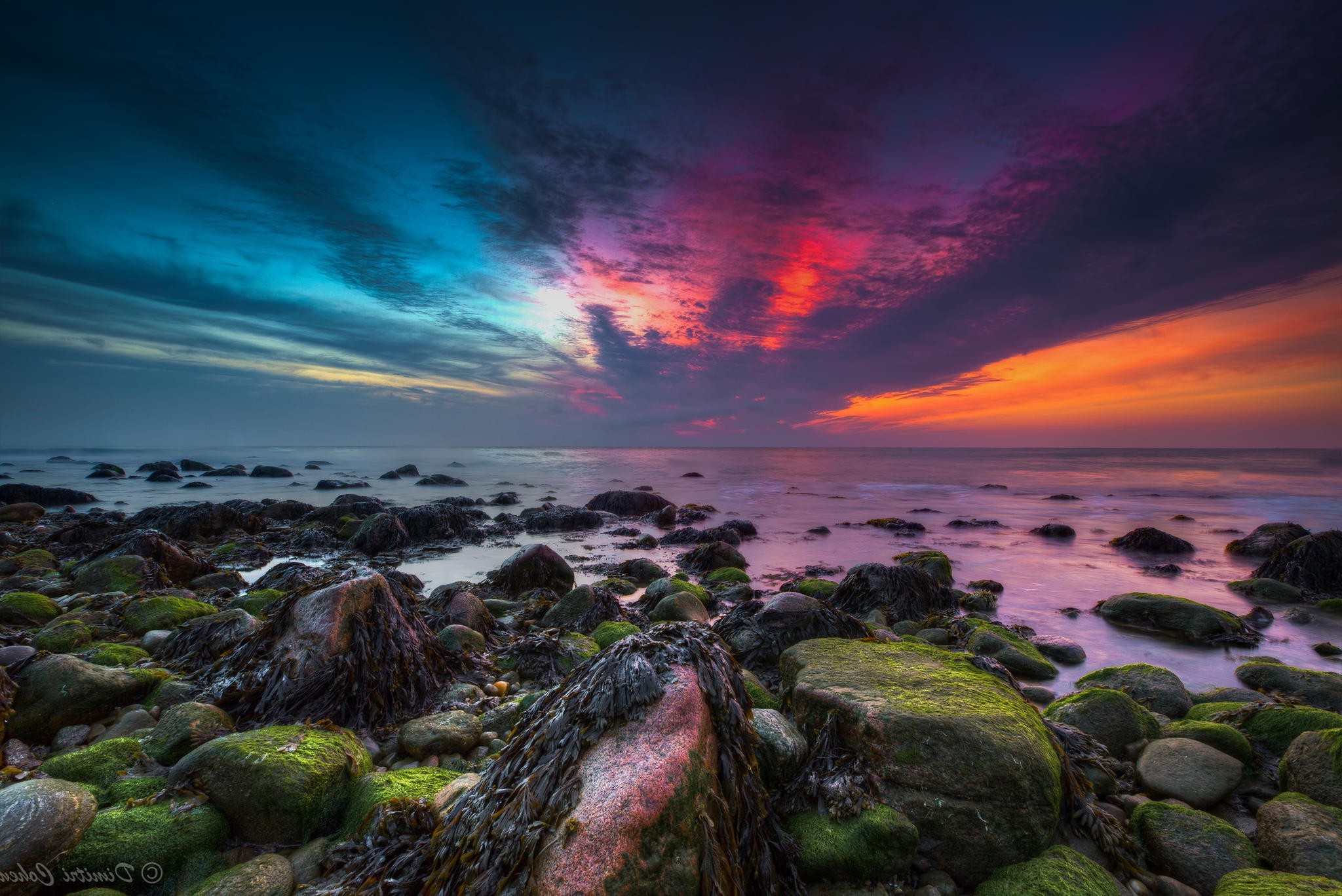 Vivid sunset over a rocky coastline, with the sky painted in shades of orange, pink, and teal, and the rocks covered in moss and seaweed
