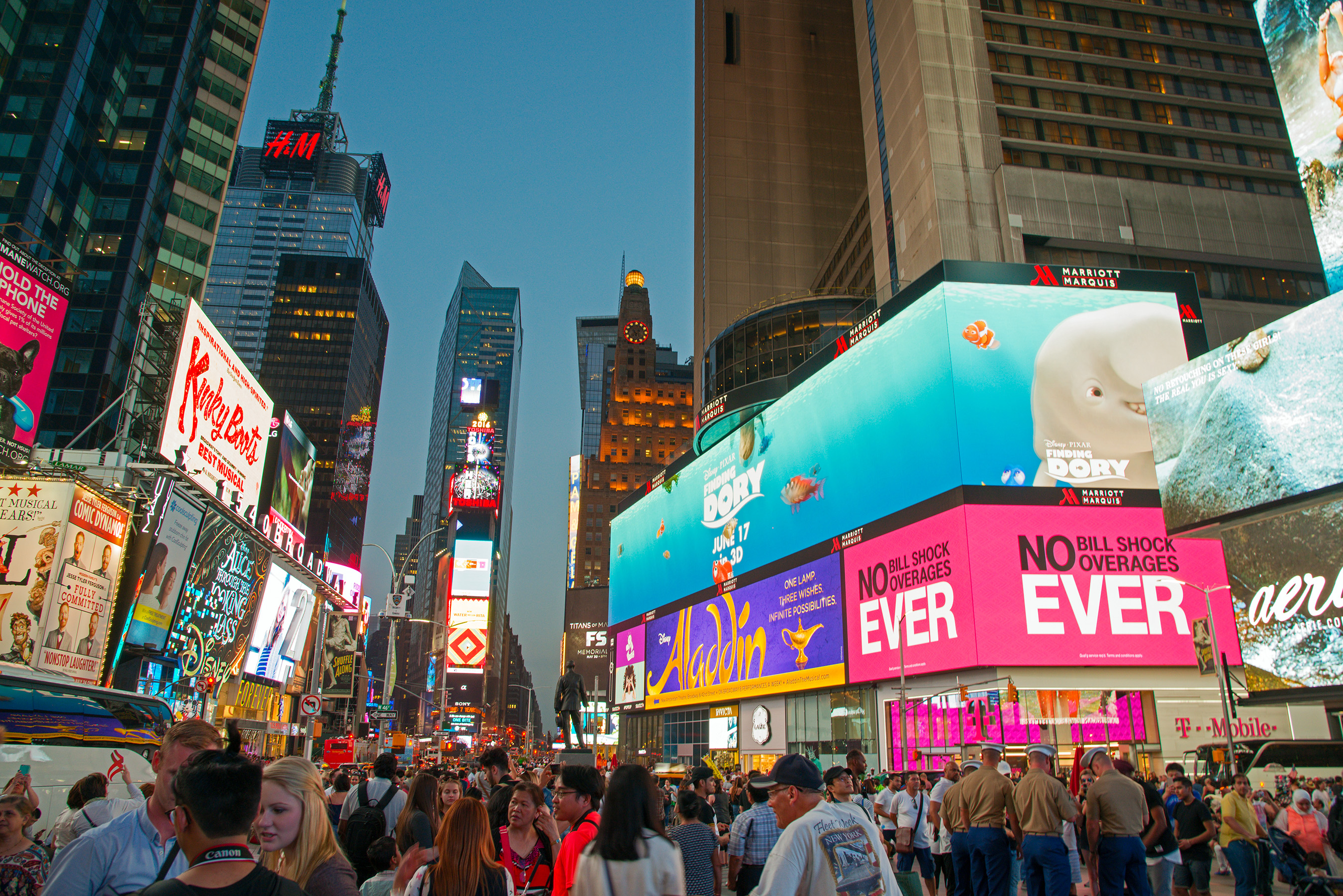 A vibrant night scene in Times Square, showcasing the bright lights and bustling crowds of Broadway, with prominent advertisements for shows like 'Kinky Boots', 'Aladdin', and 'Finding Dory'