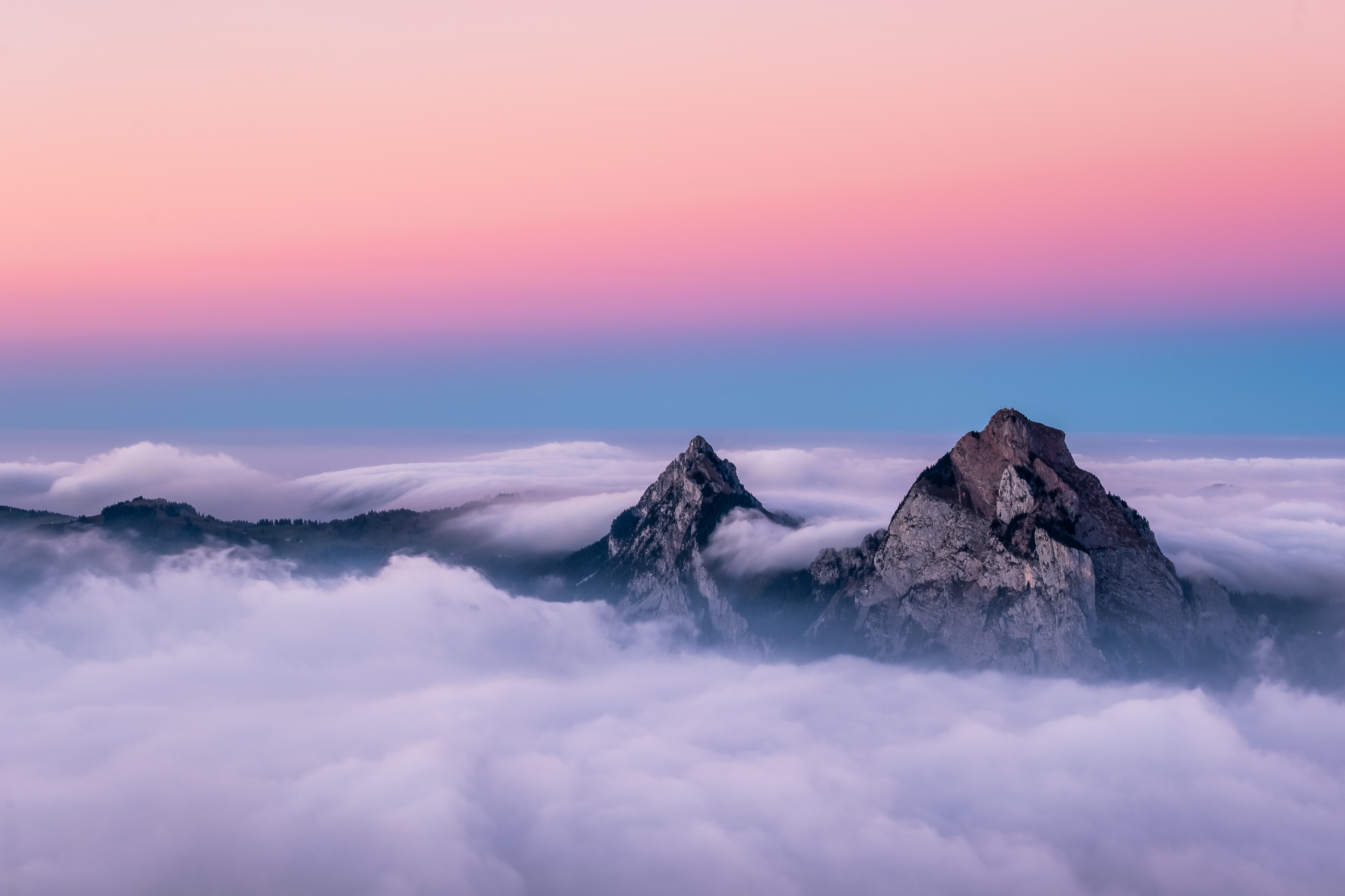 Two majestic mountain peaks rise above a sea of clouds, bathed in the soft, pastel hues of a breathtaking sunset. This stunning 4K wallpaper captures the serene beauty and awe-inspiring grandeur of nature