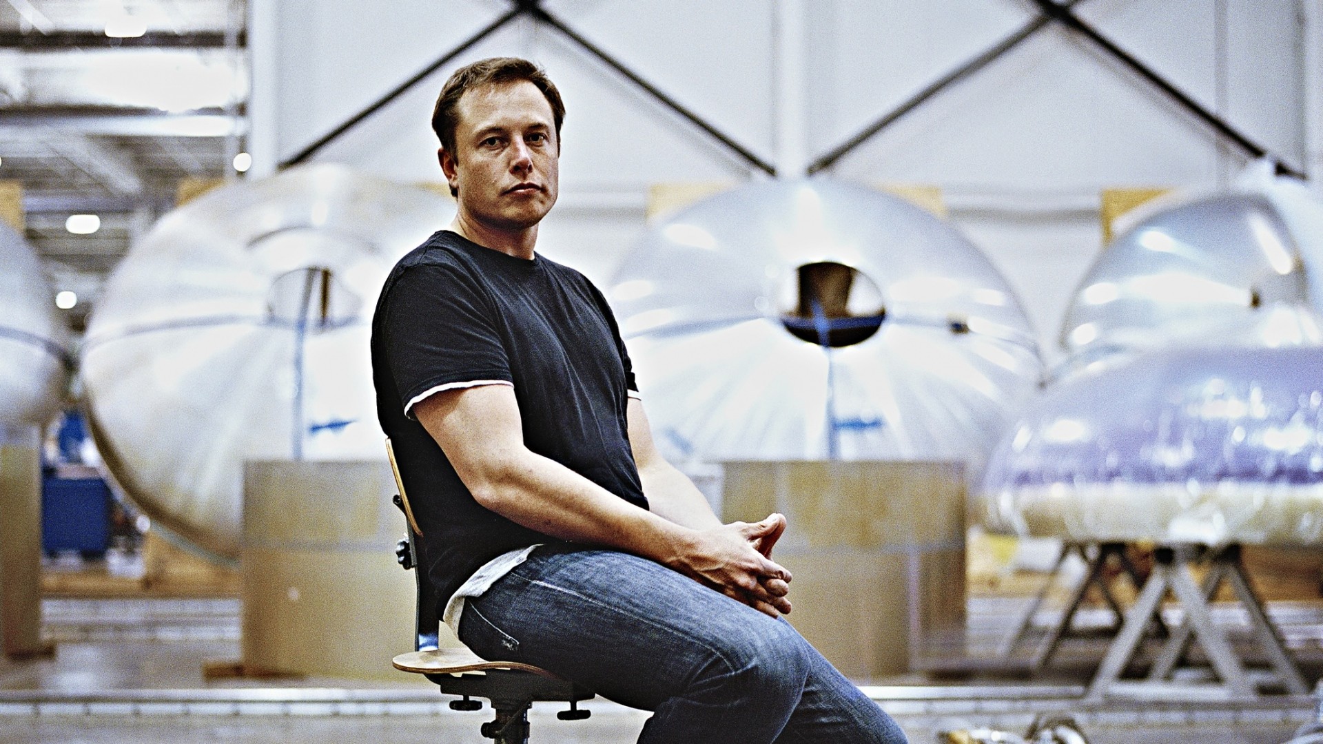 Elon Musk sitting in a chair in a SpaceX factory with Starship components in the background