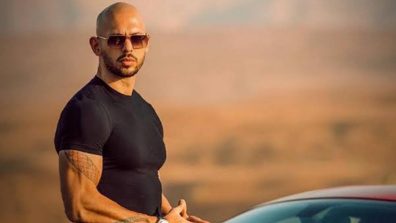 Andrew Tate posing in front of a blurred background, wearing a black t-shirt and sunglasses
