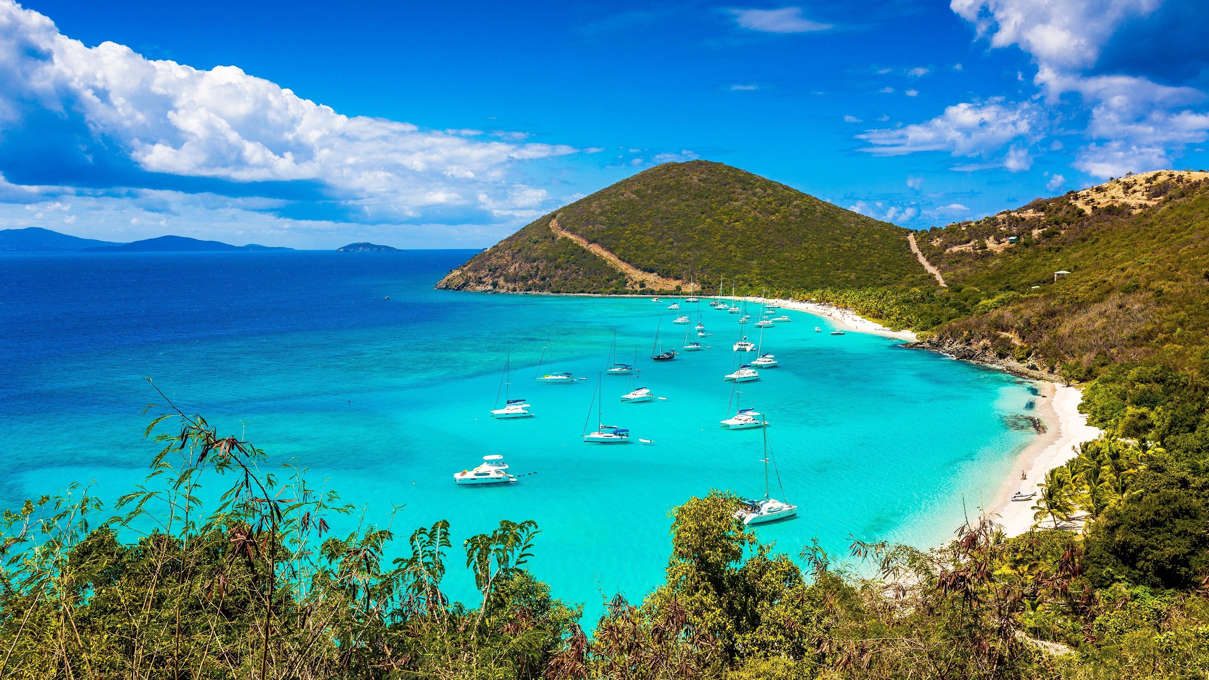 Scenic view of a tropical island bay with turquoise water, white sand beach, lush greenery, and sailboats anchored in the calm waters