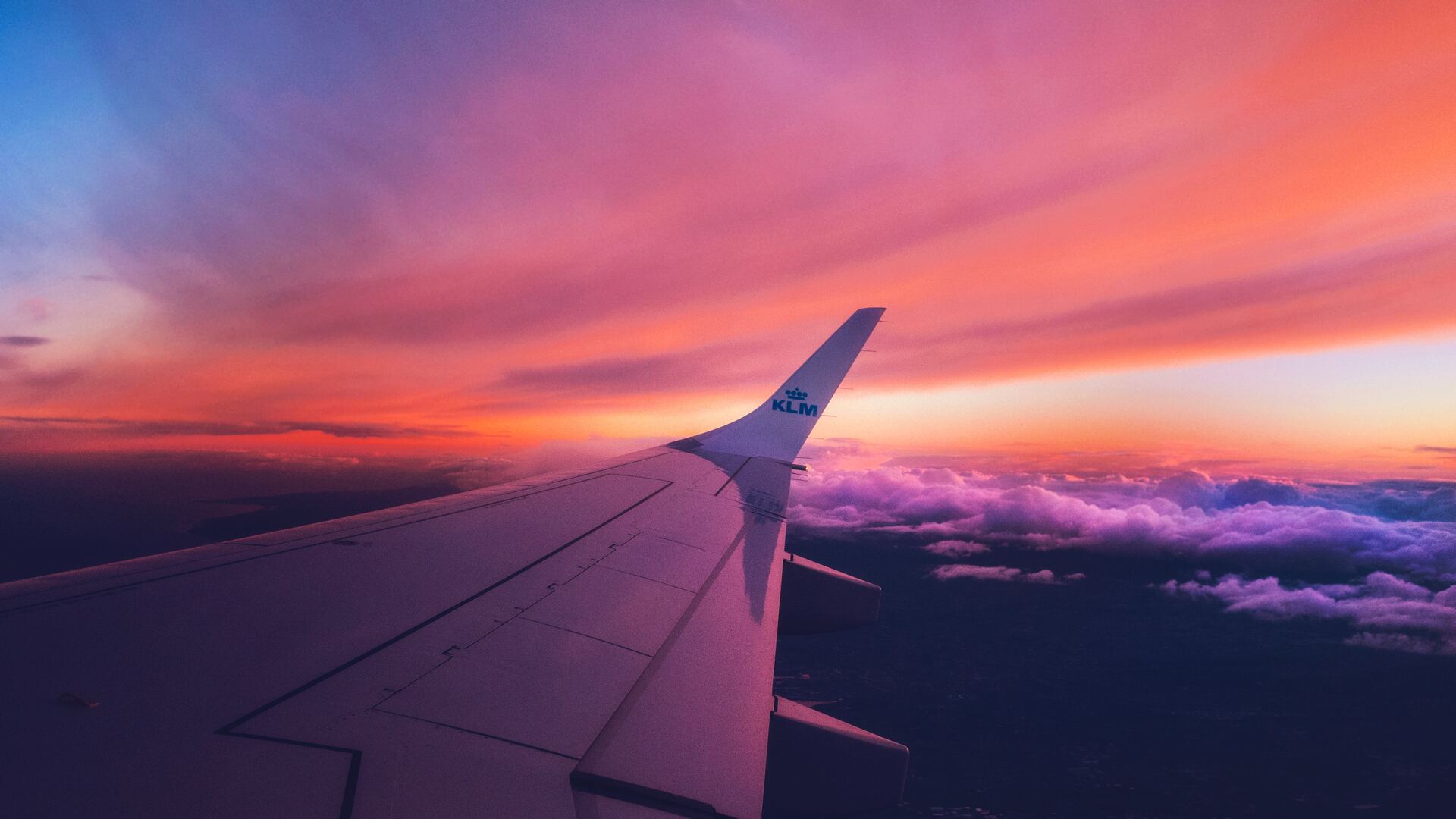 Airplane wing soaring through a vibrant sunset sky with a layer of purple-tinged clouds below, offering stunning plane backgrounds