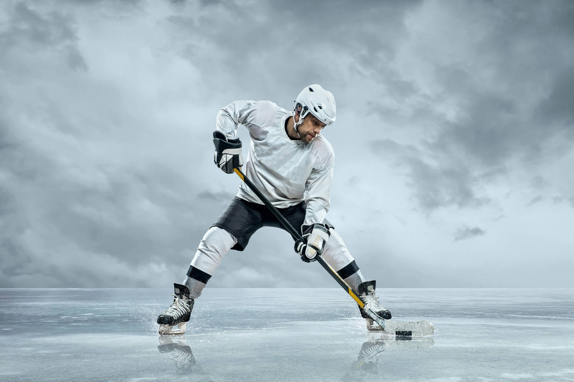 A hockey player in a white jersey and black shorts aggressively handles the puck on an ice rink beneath a stormy sky