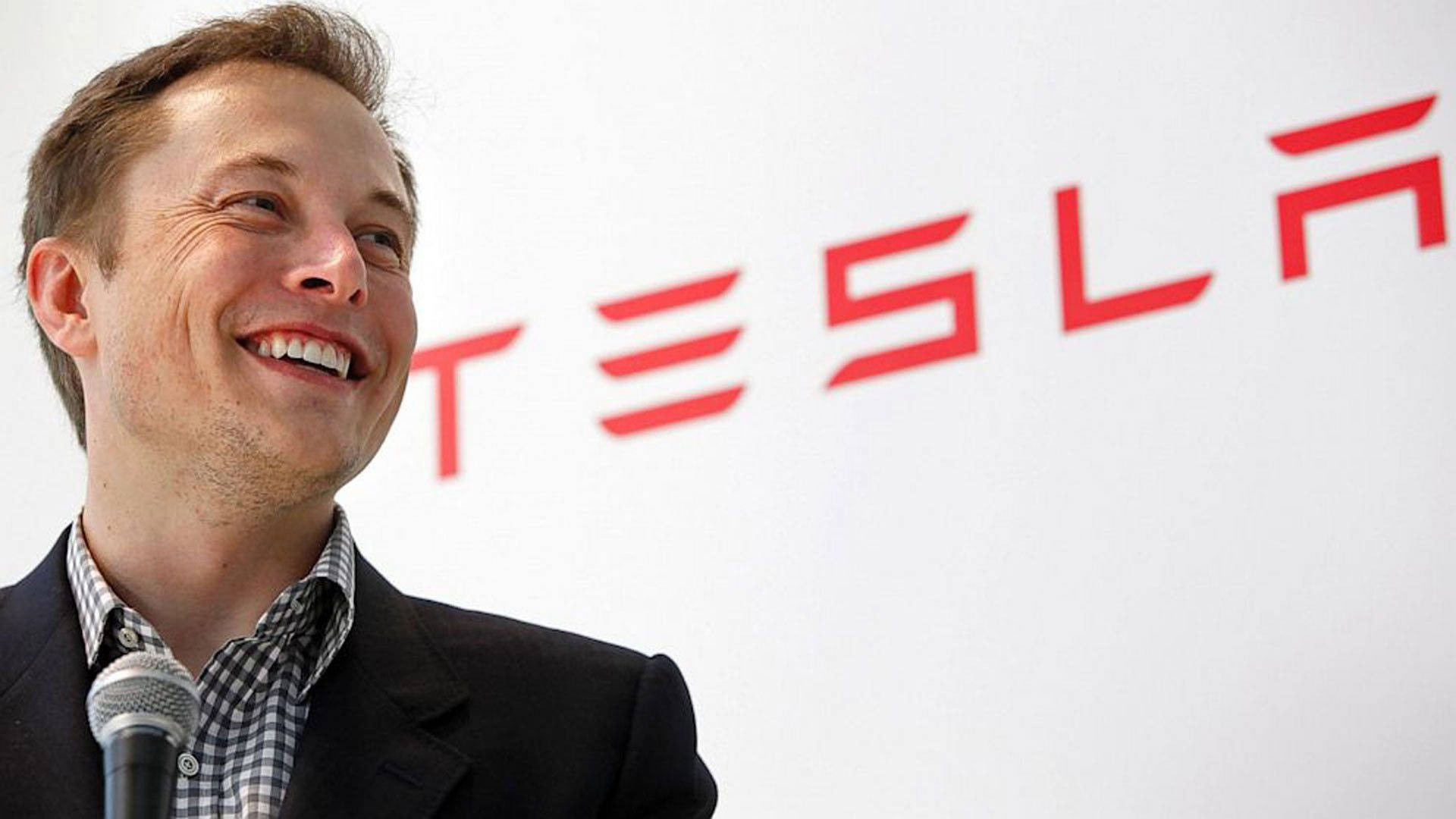 Elon Musk, CEO of Tesla, smiles at an event with the Tesla logo in the background