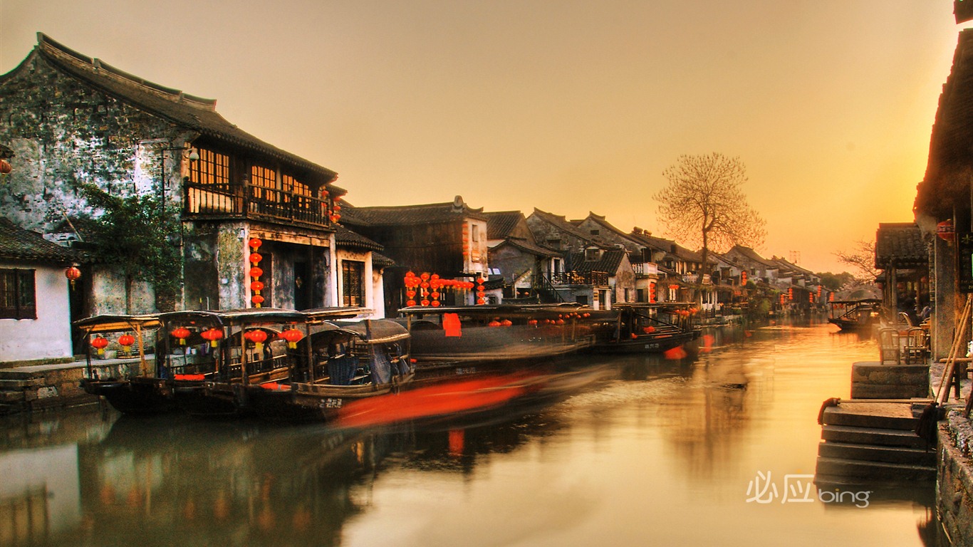 A picturesque canal in a traditional Chinese town at sunset, adorned with red lanterns and boats