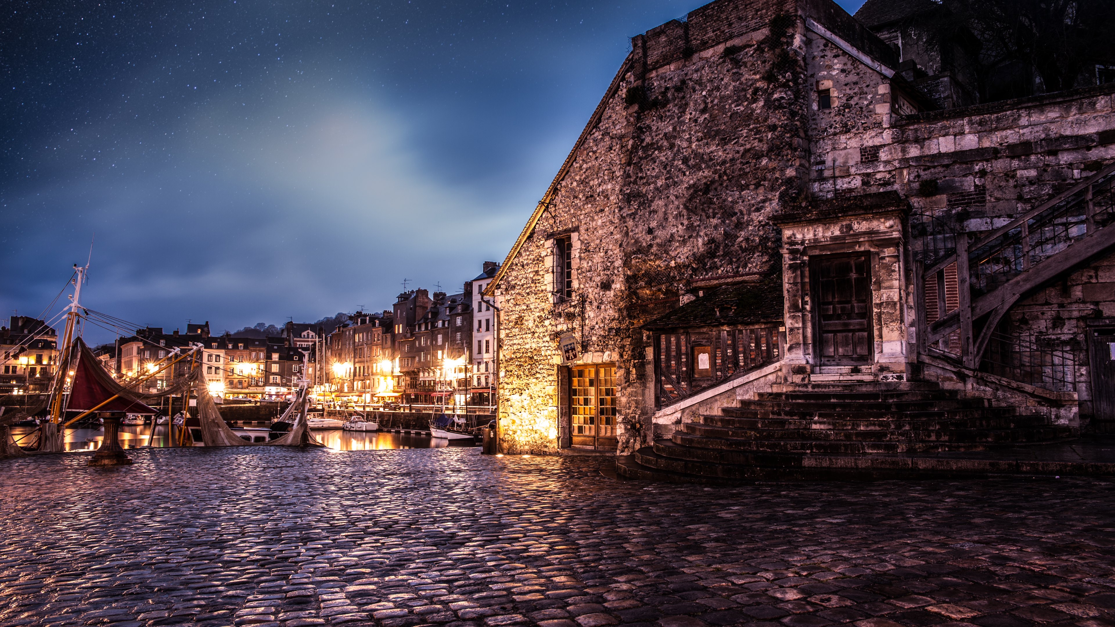 A cobblestone quay at night, illuminated by the warm glow of lights from buildings and boats, under a starry sky