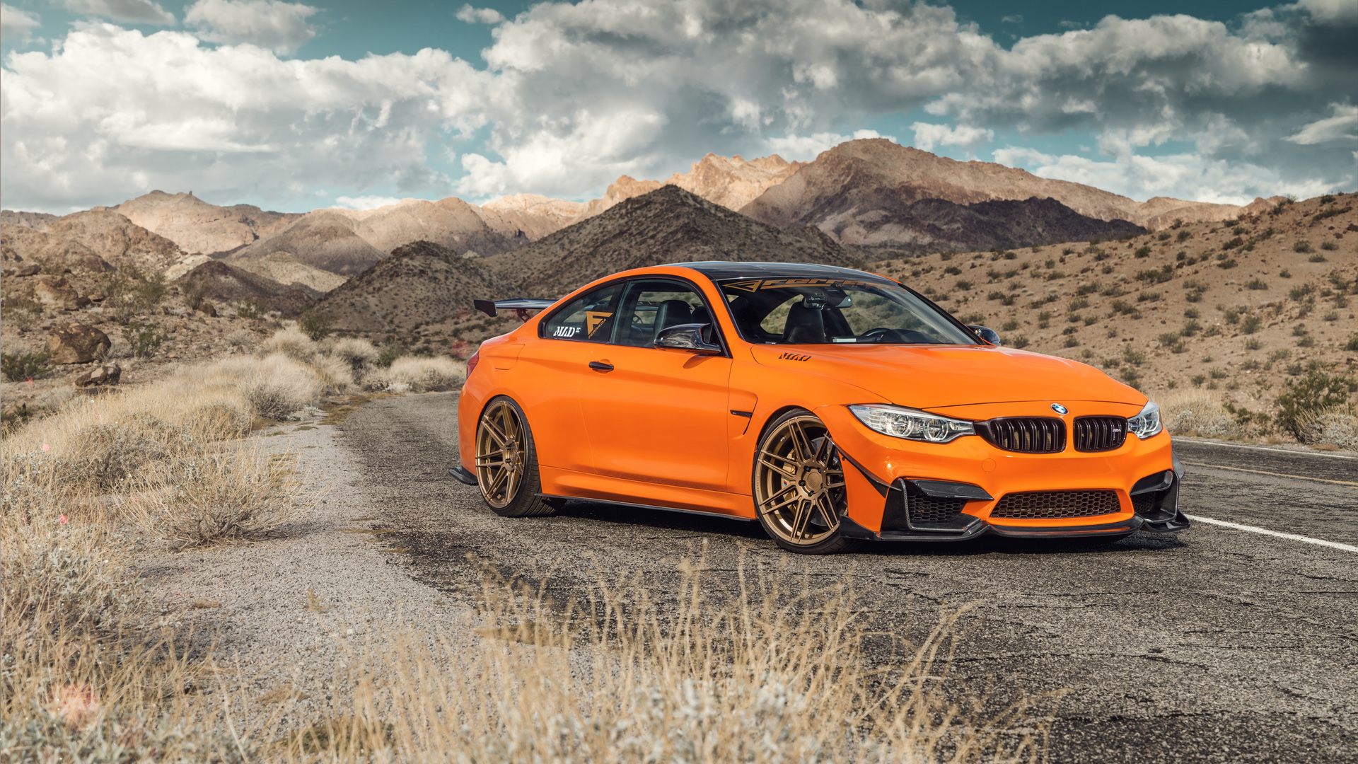 A vibrant orange BMW M4 coupe parked on a desert road, with rugged mountains in the background