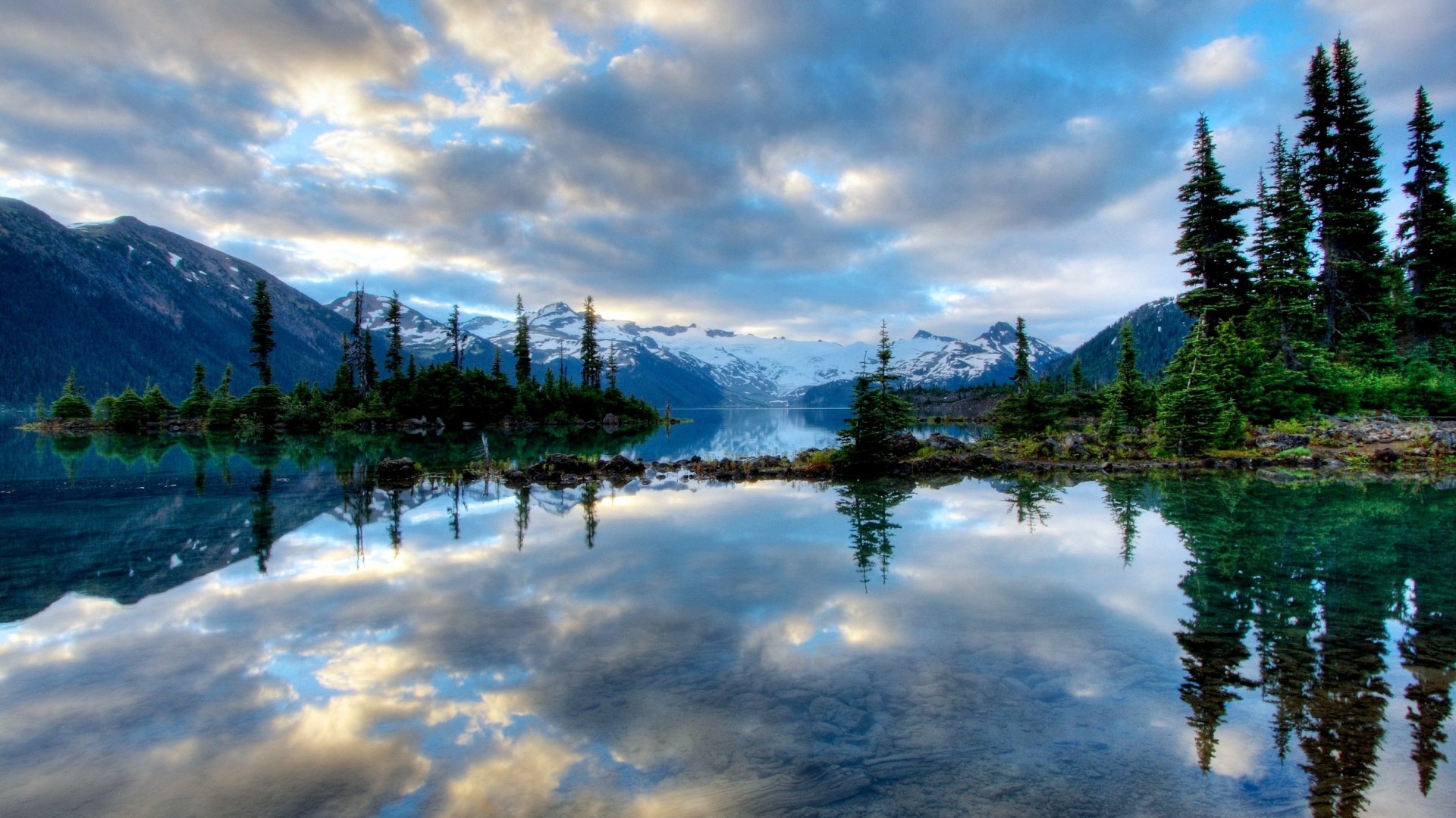 Serene mountain lake reflecting the sky and clouds, with evergreen trees on the shore and snow-capped mountains in the background