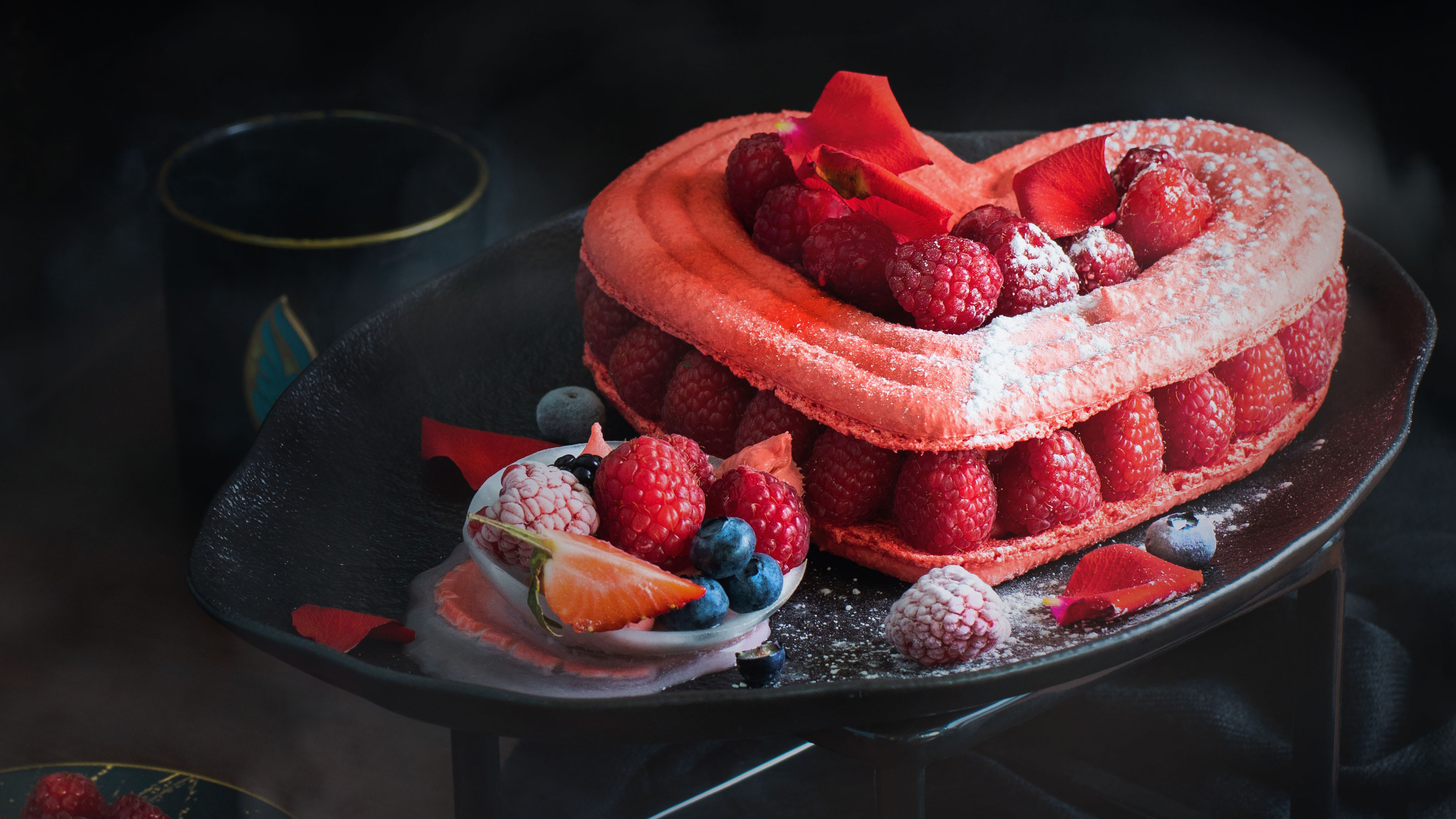 Heart-shaped dessert made with pink macarons, filled with fresh raspberries and topped with powdered sugar, rose petals, and fresh berries