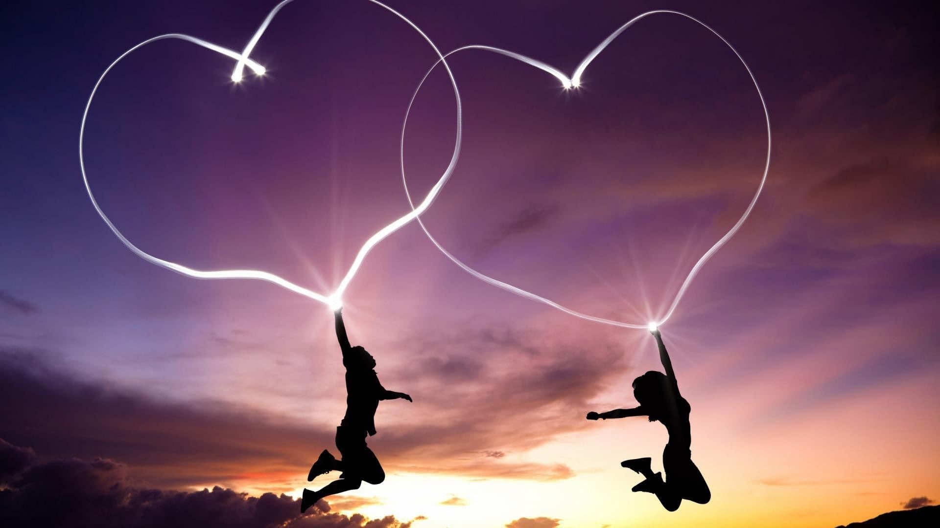 Silhouettes of two people jumping joyfully while holding onto illuminated heart shapes against a vibrant sunset sky