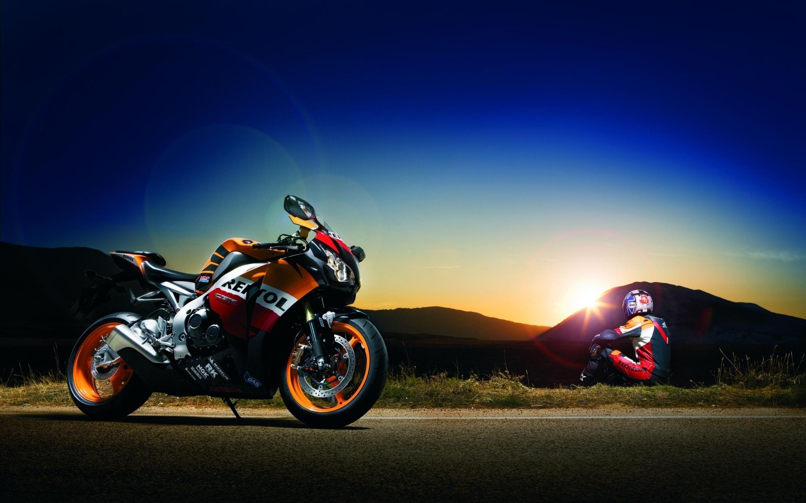 A Repsol Honda CBR1000RR Fireblade sportbike parked on a roadside at sunset with a rider admiring the view