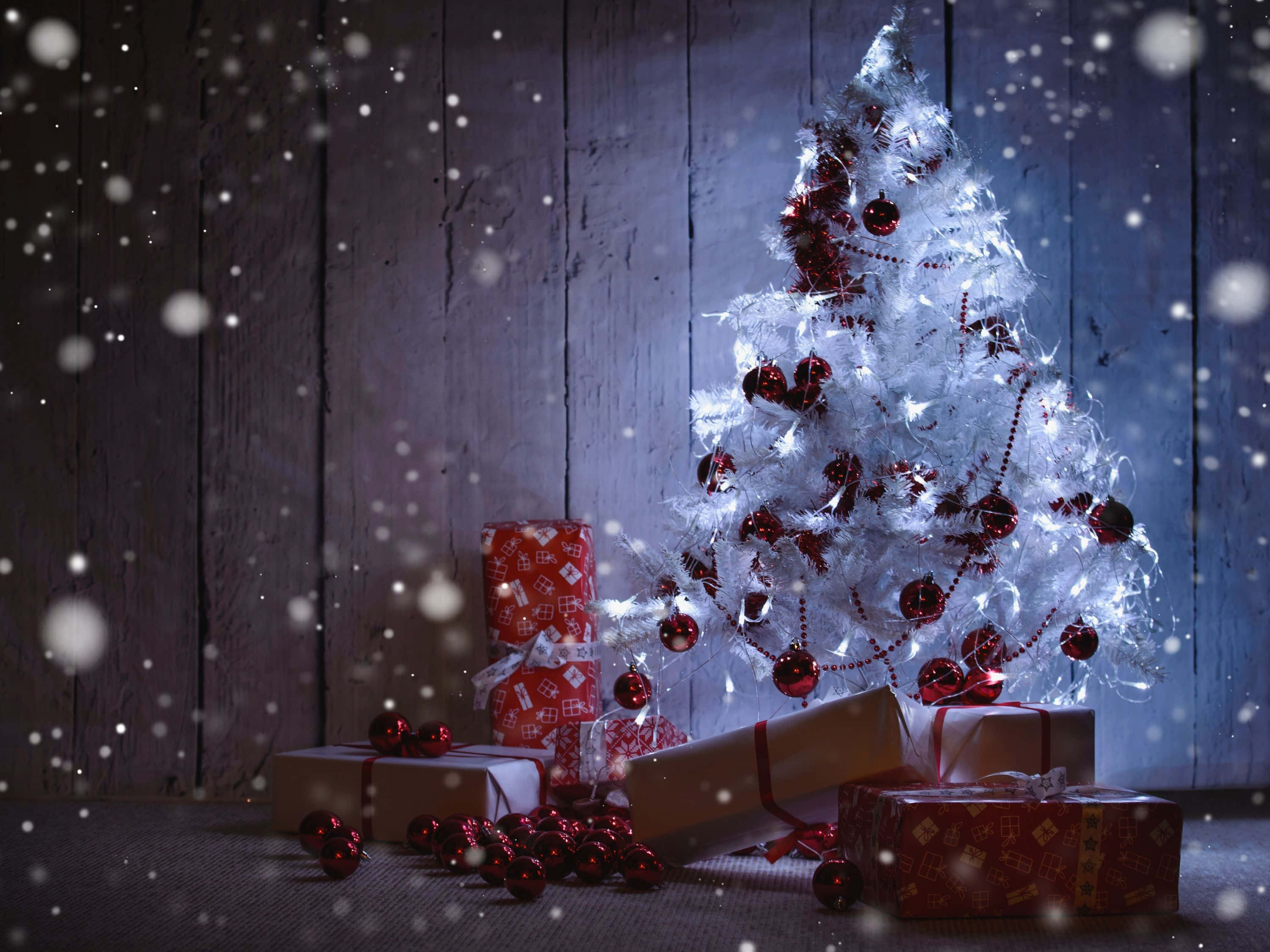 A white Christmas tree adorned with red ornaments and twinkling lights, surrounded by wrapped gifts against a rustic wooden backdrop with a gentle snowfall effect