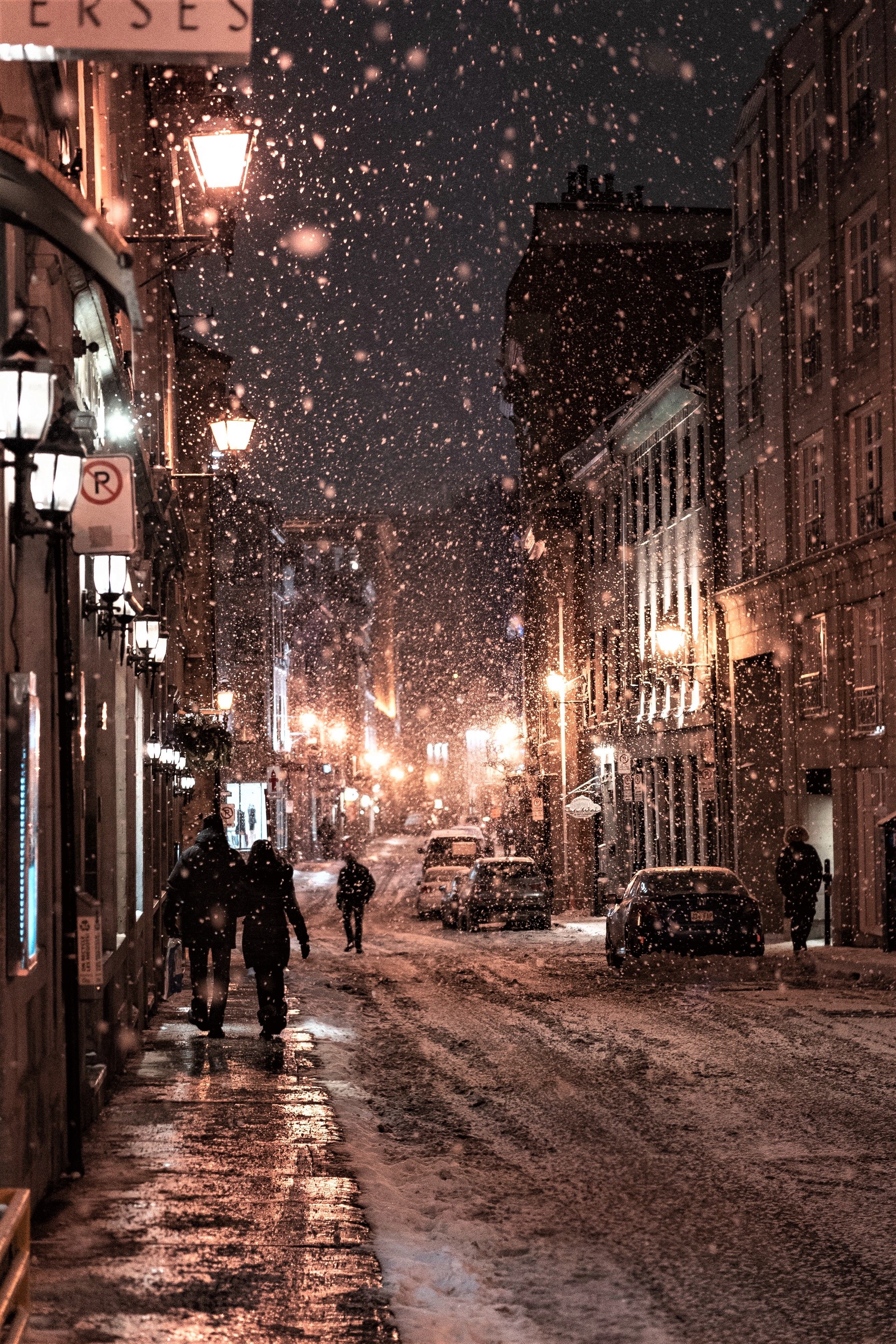 A cozy city street at night during a snowfall. Warm streetlights illuminate the snow-covered road and sidewalk as people walk by