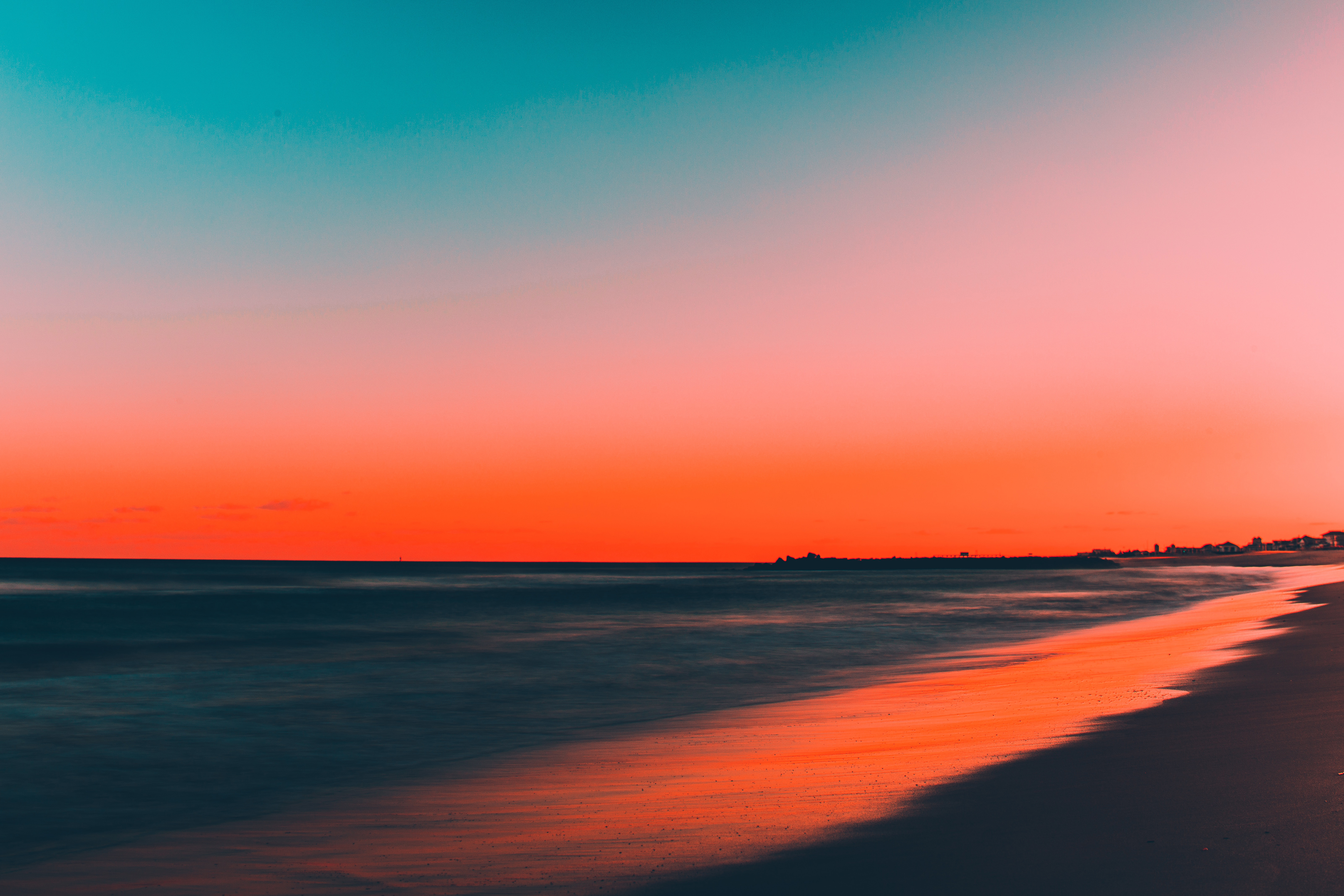 Vivid sunset over a tranquil beach, with the sky ablaze in vibrant hues of orange, pink, and teal. The gentle waves create a soft blur, adding to the serene and picturesque scene