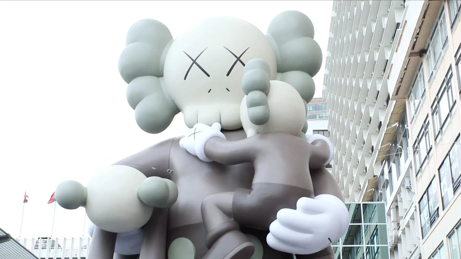 A large inflatable Kaws Companion figure holding a smaller Companion figure, displayed outdoors against a backdrop of city buildings