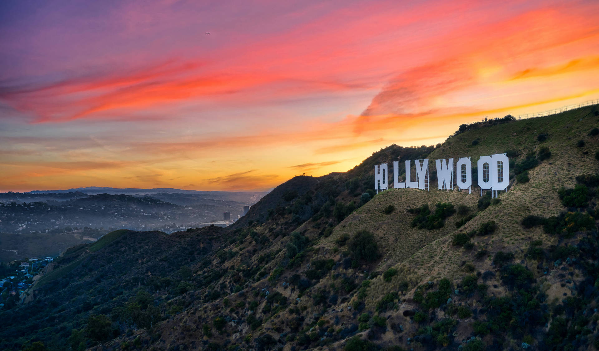 The iconic Hollywood Sign at sunset, overlooking the Los Angeles cityscape. Vibrant orange, pink, and purple hues paint the sky in this stunning wallpaper image