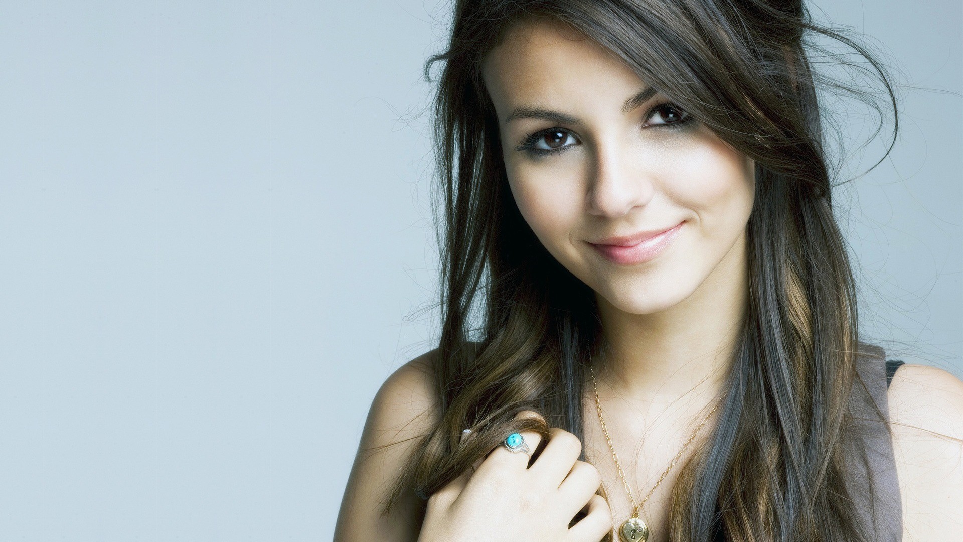 A beautiful young woman with long brown hair smiles gently at the camera. She's wearing a delicate gold necklace and a turquoise ring, and her expressive eyes captivate the viewer