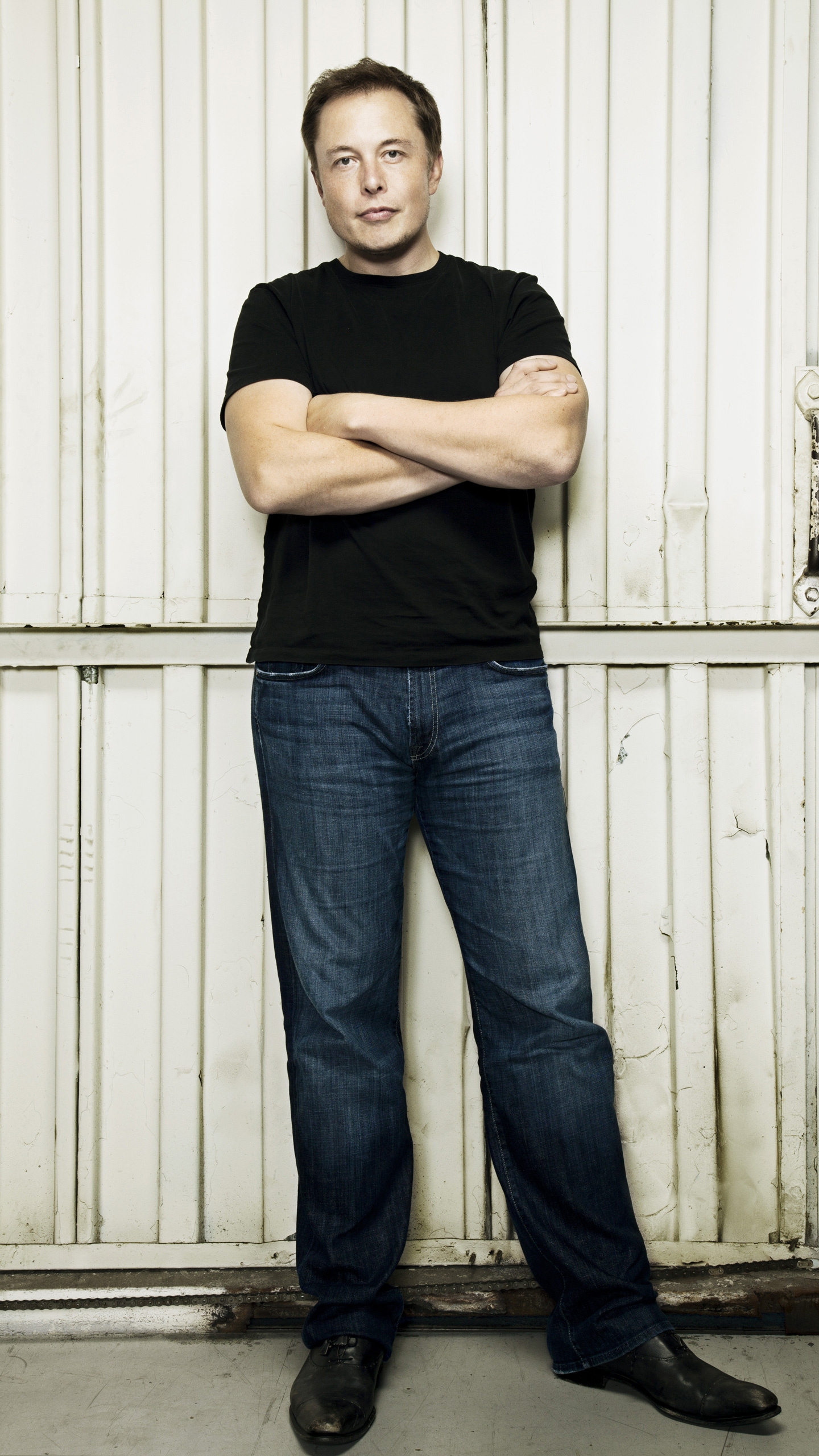Elon Musk standing against a white wall with his arms crossed, wearing a black t-shirt and jeans