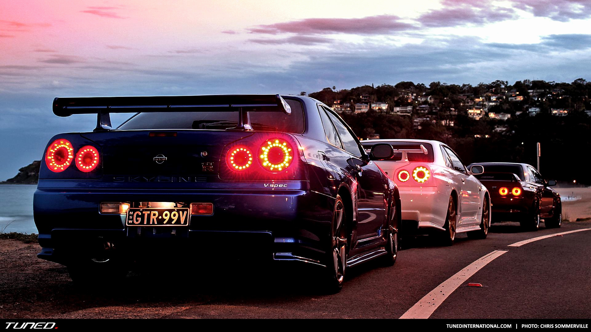 A lineup of Nissan Skyline GT-Rs, reminiscent of the Fast and Furious franchise, parked along a scenic coastal road at dusk