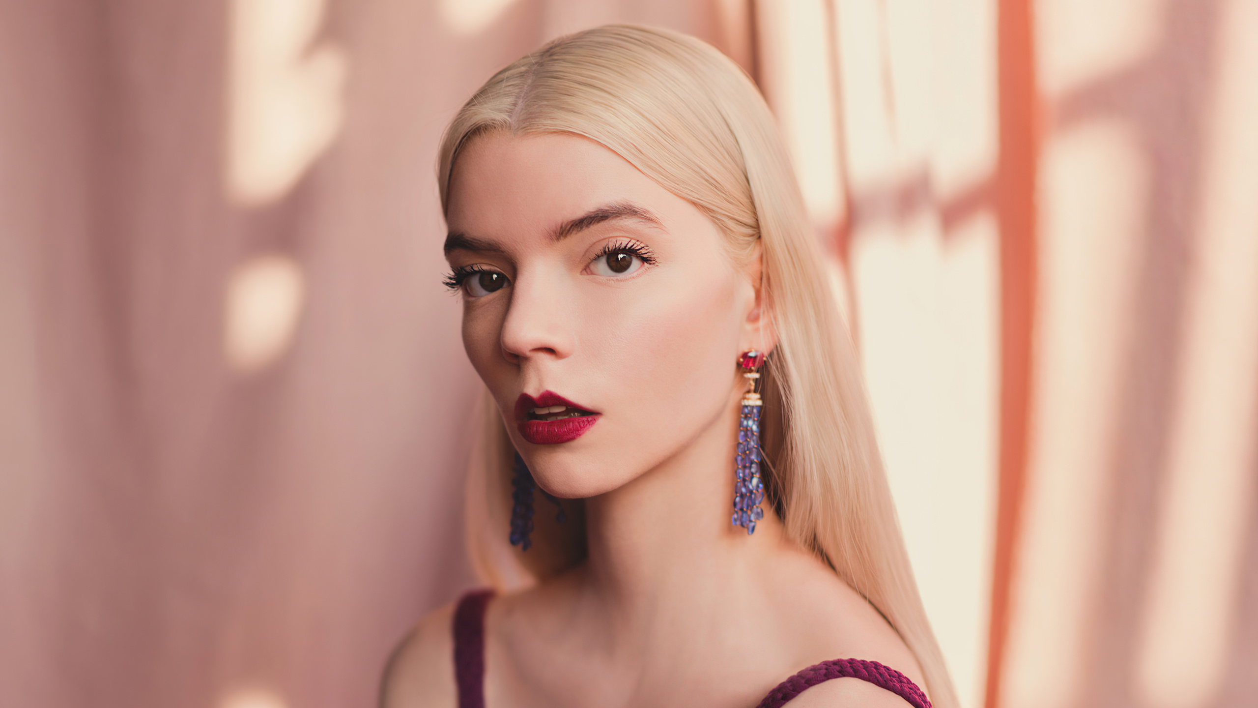 Anya Taylor-Joy posing in front of a pink background, wearing a maroon top and purple tassel earrings