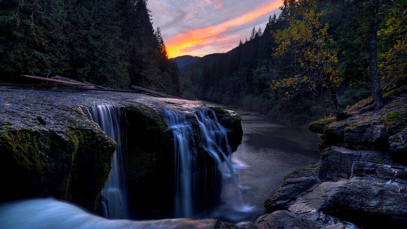 A breathtaking waterfall cascades over mossy rocks at sunset in a serene forest, creating a picturesque desktop wallpaper