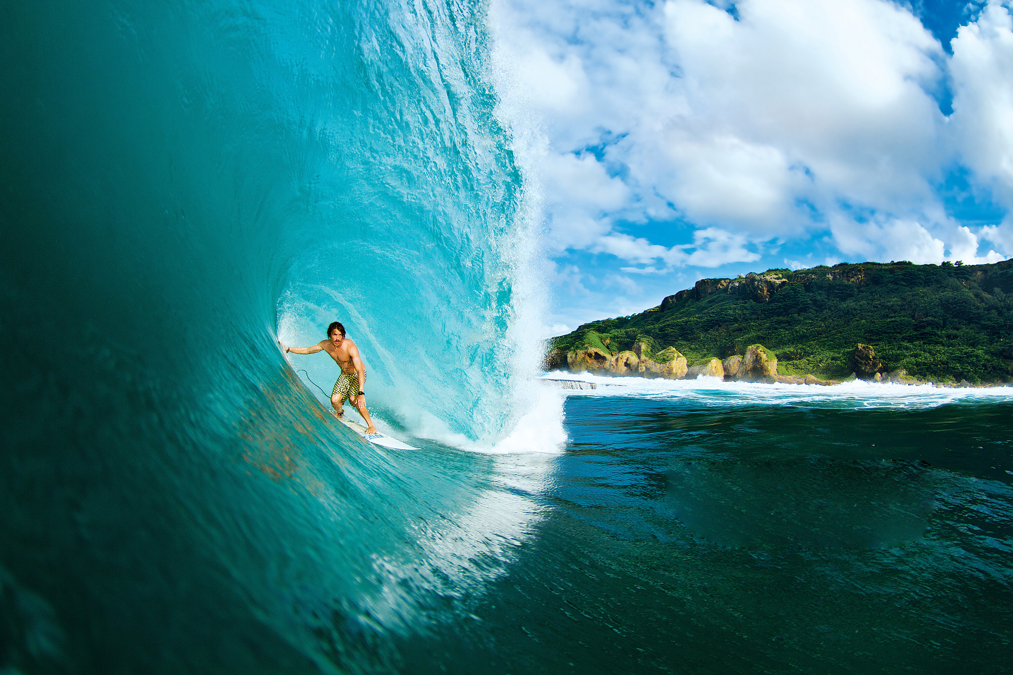 A surfer rides a powerful wave in crystal-clear turquoise water, with a lush green tropical island in the background. This dynamic surfing image captures the thrill and beauty of the sport