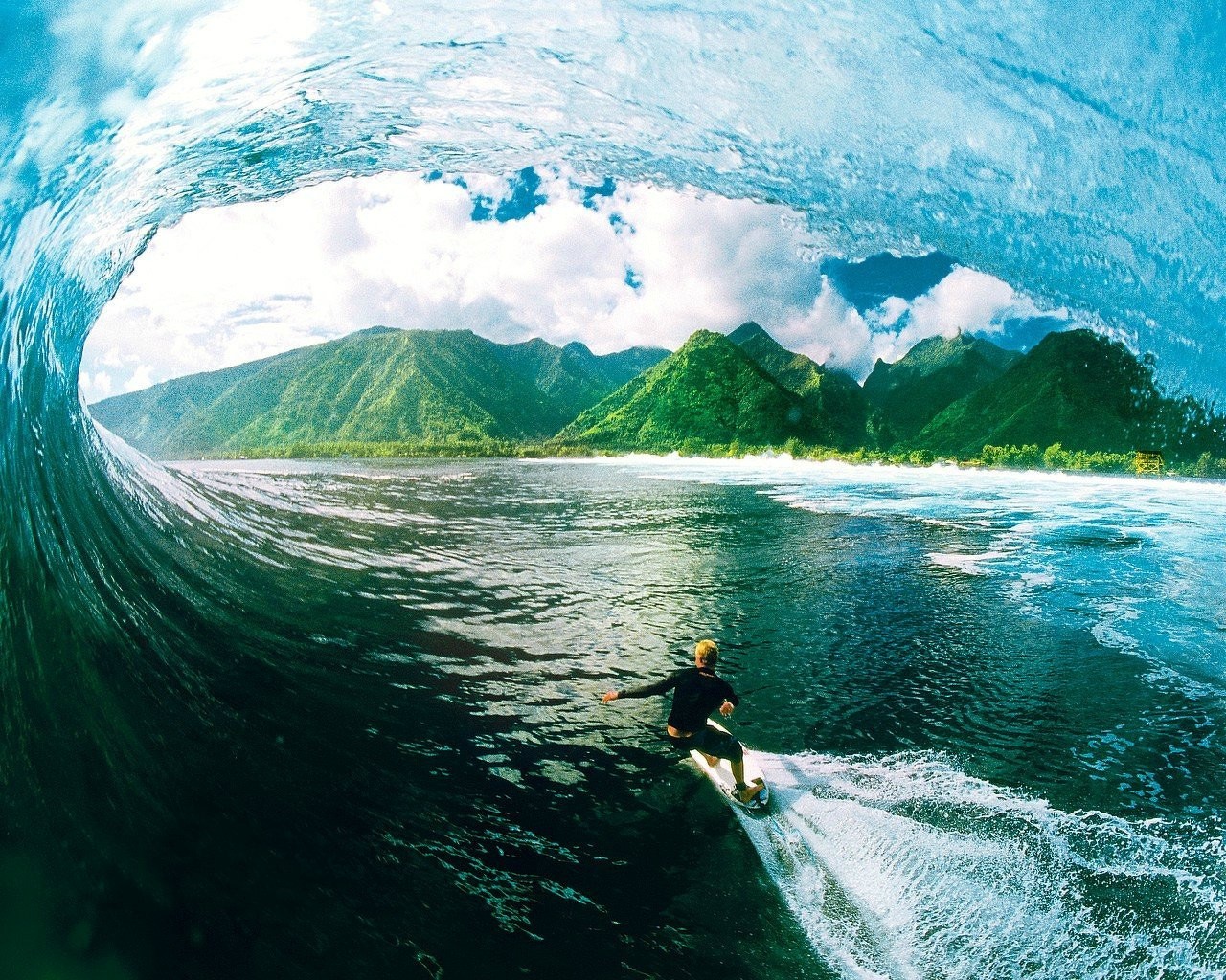 A surfer rides a massive wave in crystal-clear water, with lush green mountains visible in the background