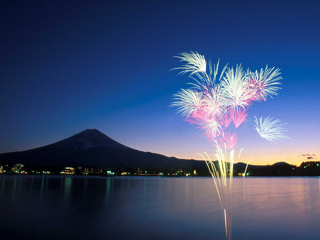 Fireworks light up the night sky over Mount Fuji and a serene lake in Japan, creating a stunning desktop wallpaper