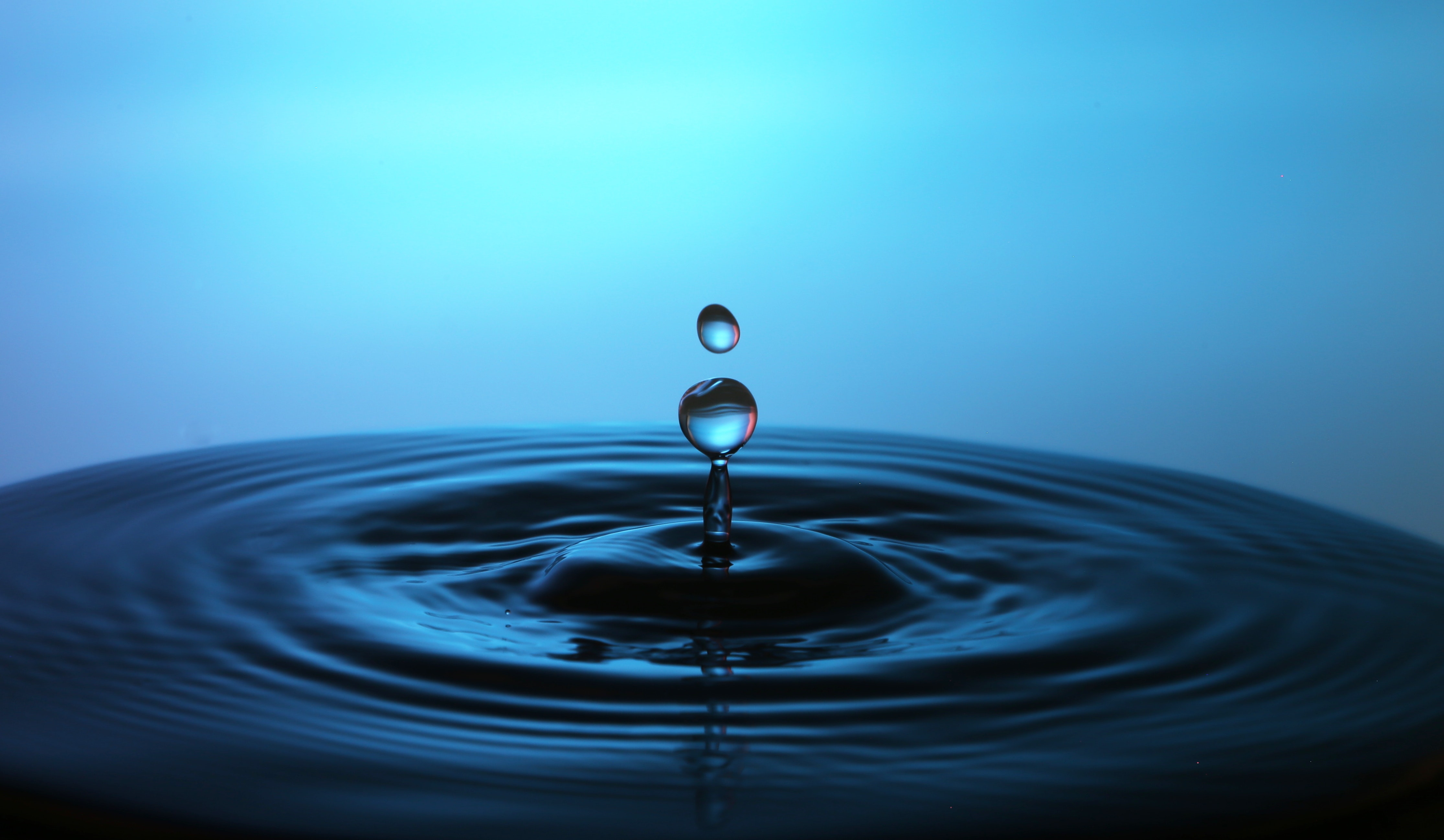 Close-up of a water droplet splashing into a pool of still water, creating concentric ripples. The background is a gradient of blue, giving a serene and refreshing feel
