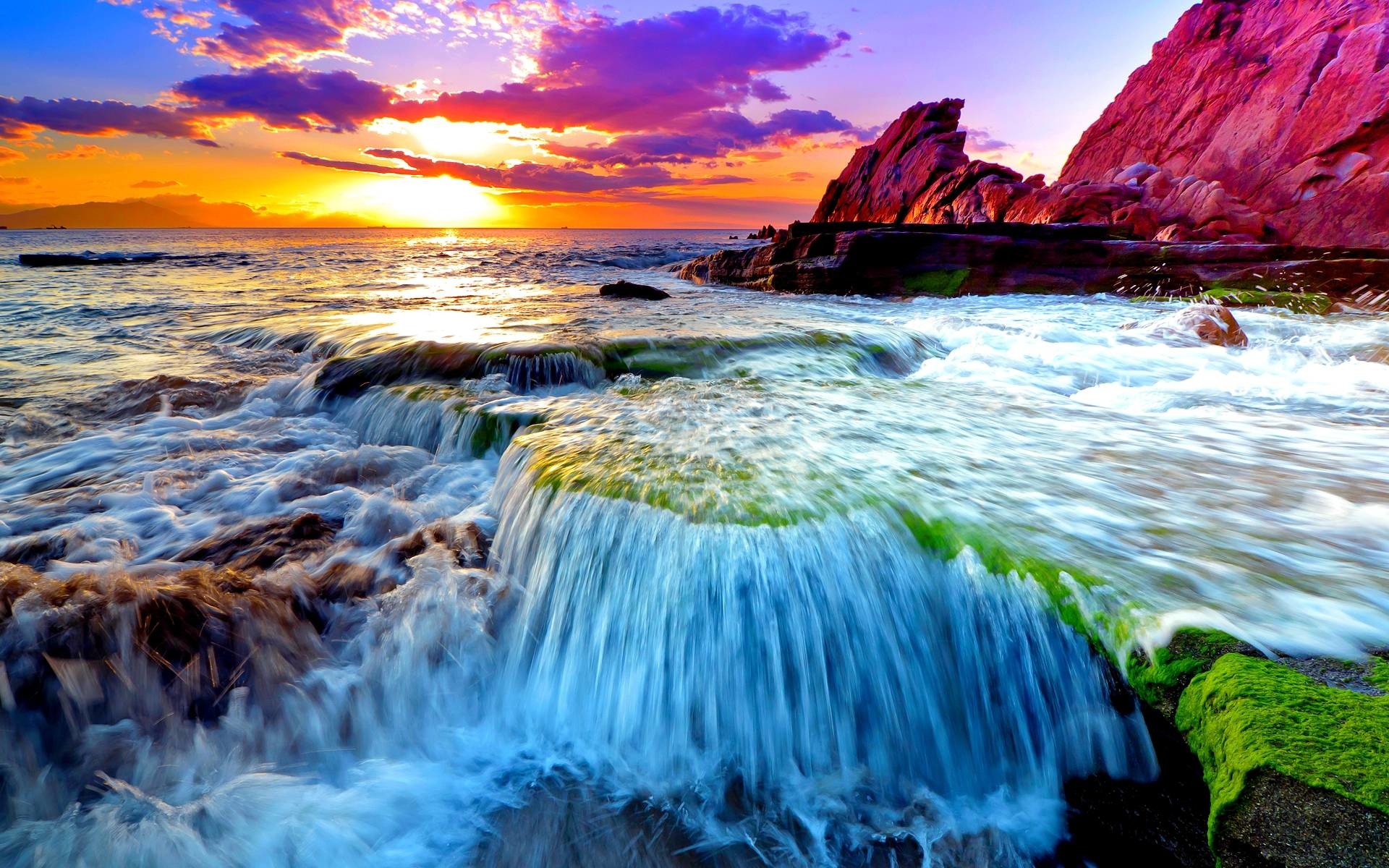 A breathtaking ocean sunset scene with waves cascading over moss-covered rocks, creating a natural waterfall effect