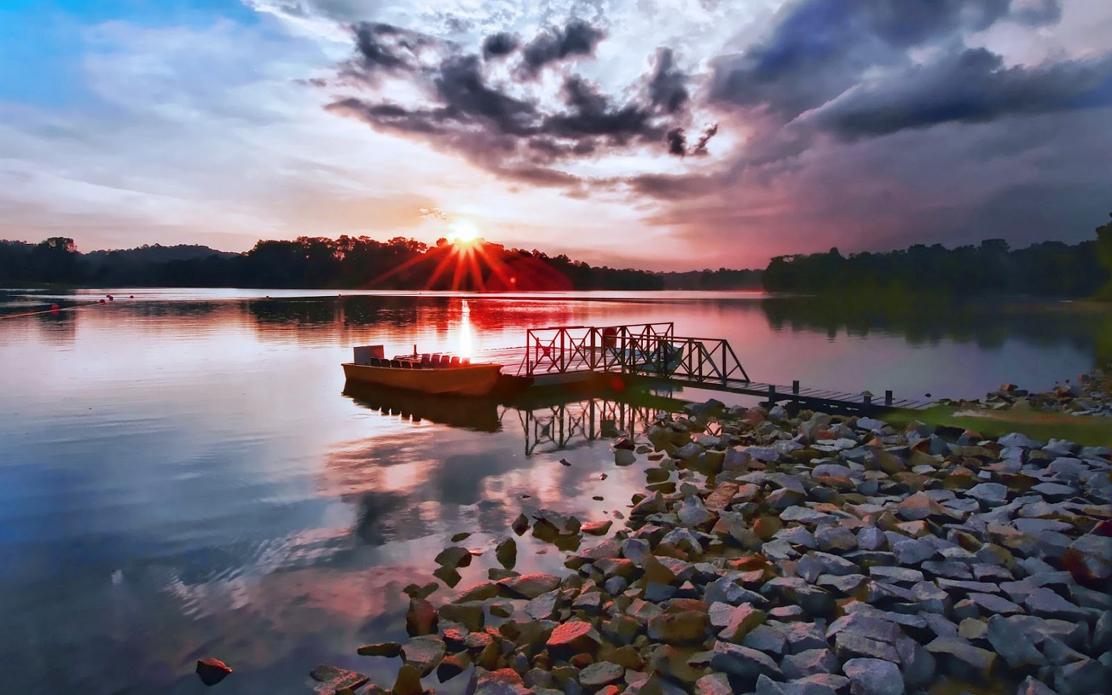 A tranquil sunset over a lake with a boat docked at a wooden pier and a rocky shoreline